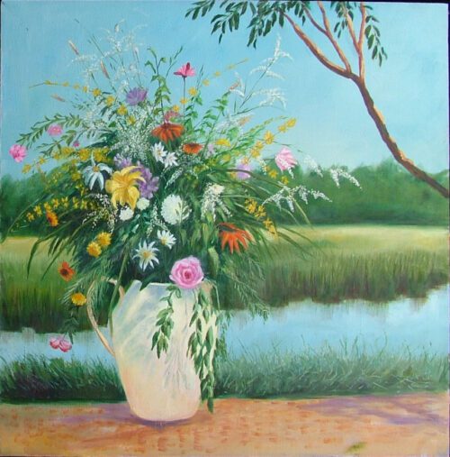 a multicolored floral bouquet is in a white vase and sits on the bank of a small river. there is green grass in the background and a tree branch hangs out of the upper right corner