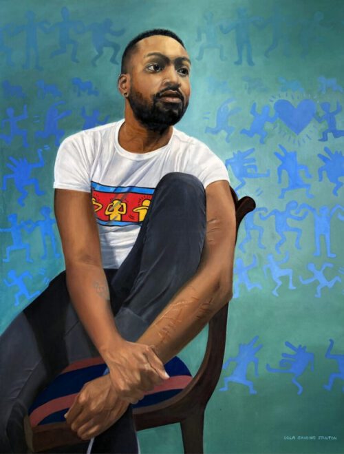 An african american man sits on a chair wearing black jeans and a white shirt with keith haring drawings. he has short hair and a beard.